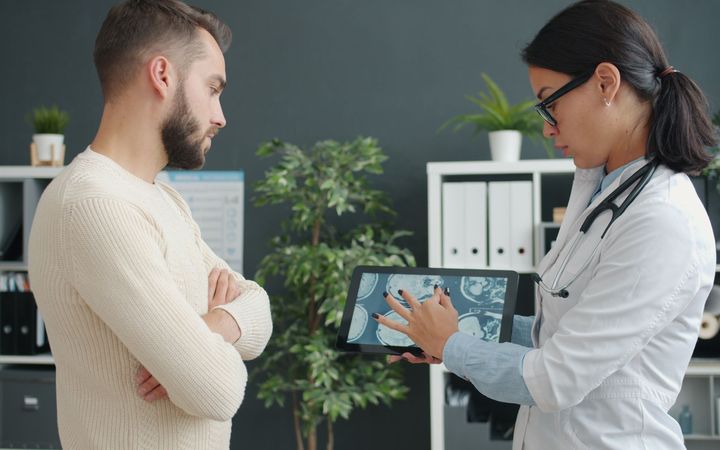 Врач показывает снимки на планшете мужчине, который стоит со скрещёнными руками