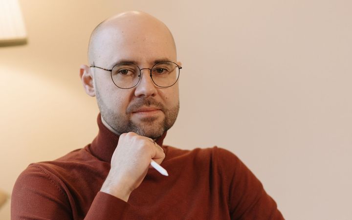Мужчина в очках и бордовом свитере сидит в кресле и задумчиво смотрит в камеру